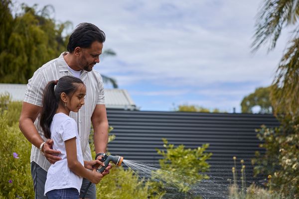 A young girl is holding the trigger of a hose pipe, while her father supports her hand and cradles her back. The couple are standing in a back garden, spraying water onto bushes.